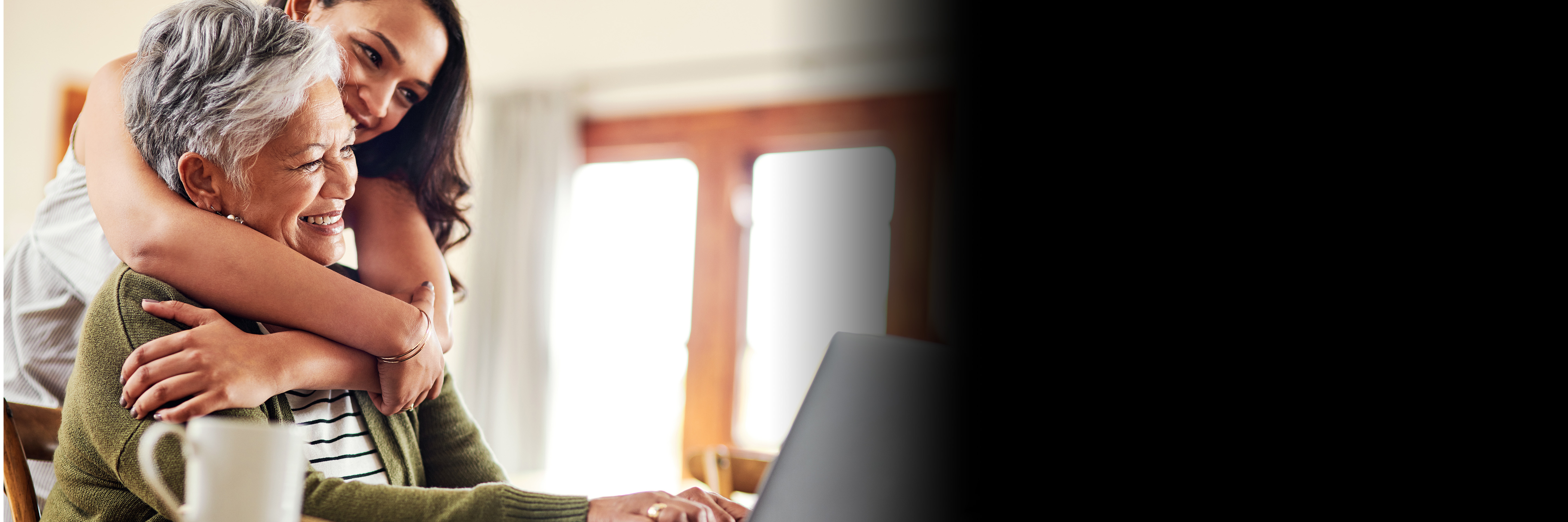 A young daughter is hugging her mother who's sitting in front of a laptop