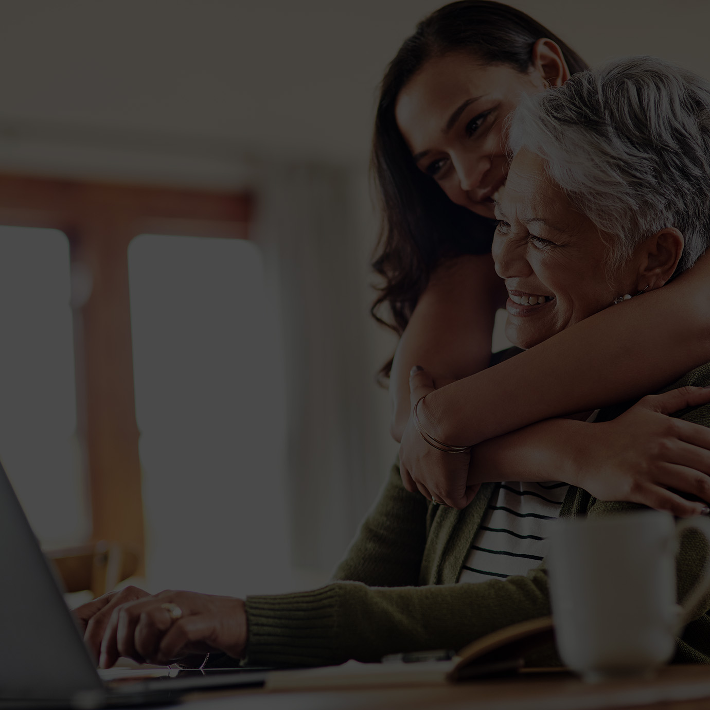 A young daughter is hugging her mother who's sitting in front of a laptop