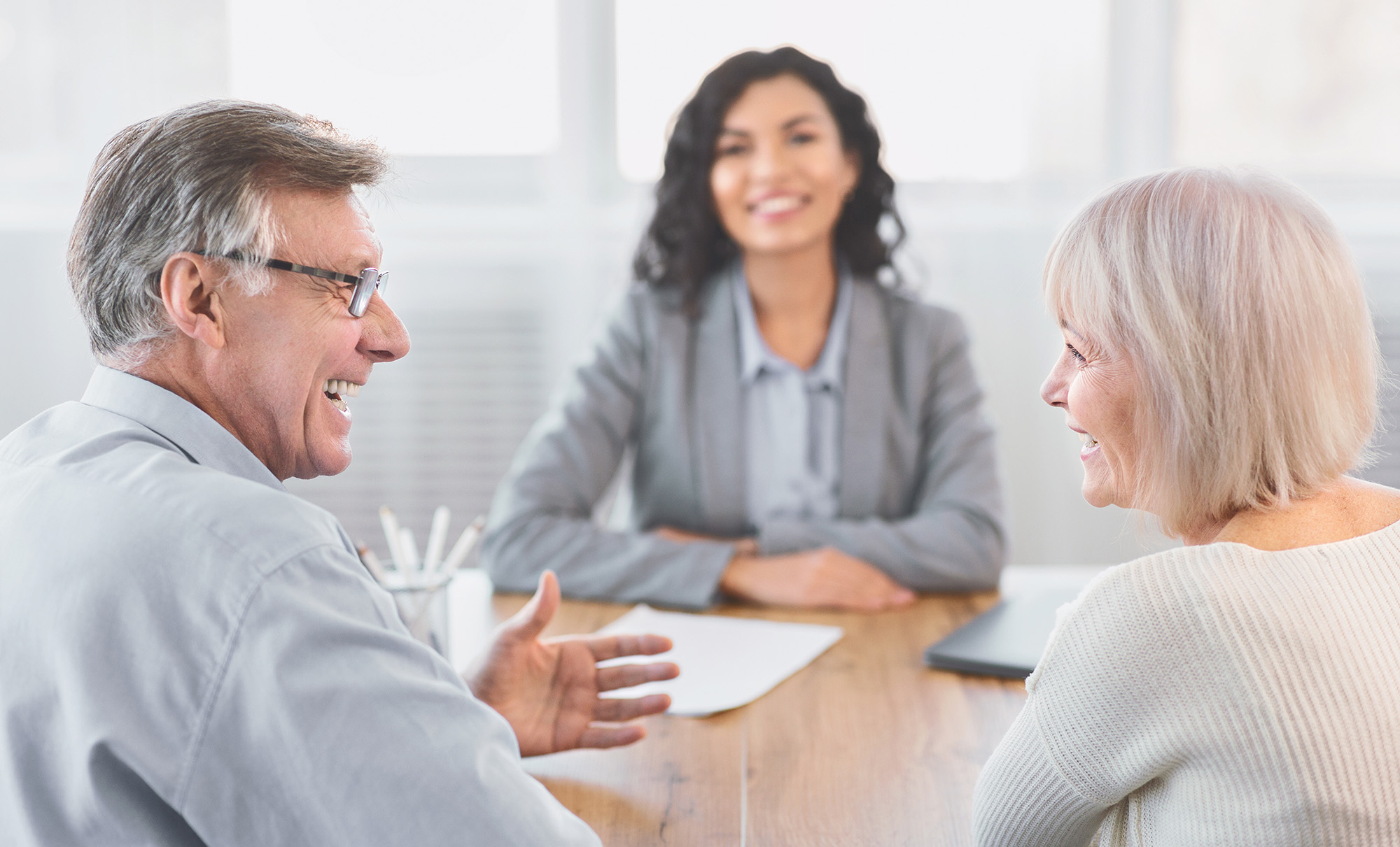 A senior couple smiling at each other with a female agent sit across the table 